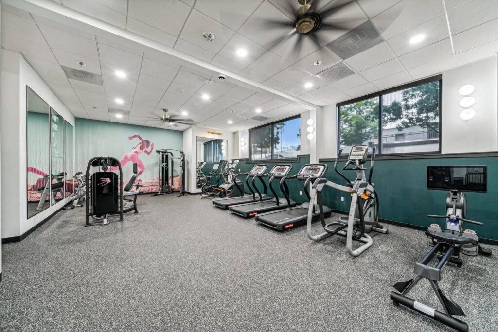 Modern gym with treadmills, elliptical machines, a stationary bike, weight machines, large mirrors, windows with outdoor views, and a wall mural of a running person. The room is bright with ceiling lights and fans.