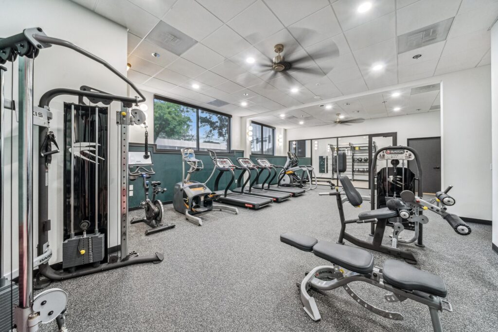 Modern gym with treadmills, exercise bikes, weight machines, benches, and free weights. Large windows provide natural light, and ceiling fans are visible. The space is clean and organized, with various workout equipment neatly arranged.