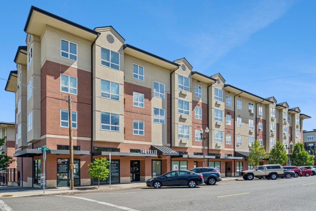 A modern four-story apartment building with tan and red brick exterior, large windows, and storefronts on the ground level. Several cars are parked along the street in front, and trees line the sidewalk under a clear blue sky.