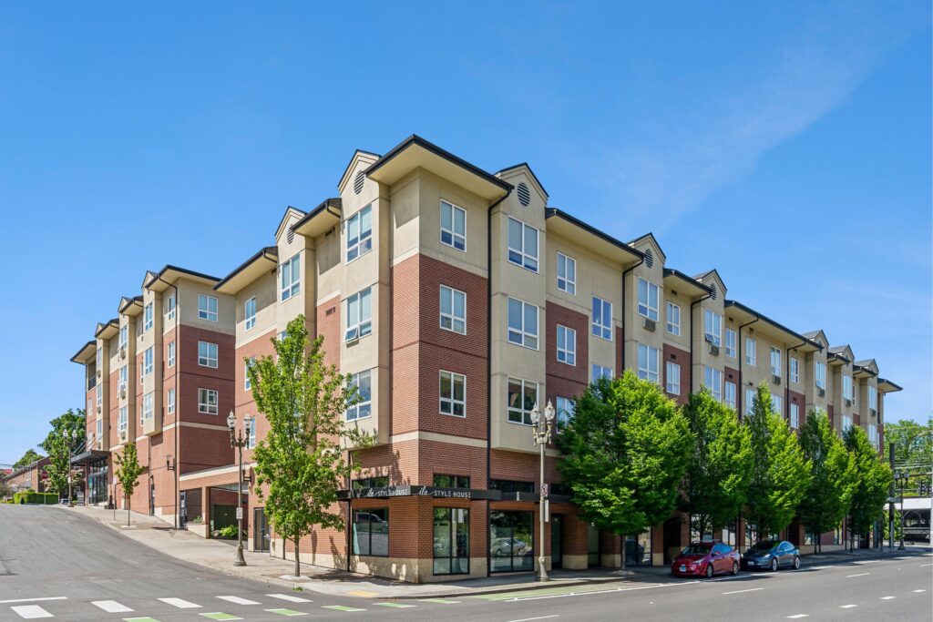 A modern four-story apartment building with ground-floor shops on a street corner, surrounded by green trees and parked cars, under a clear blue sky.