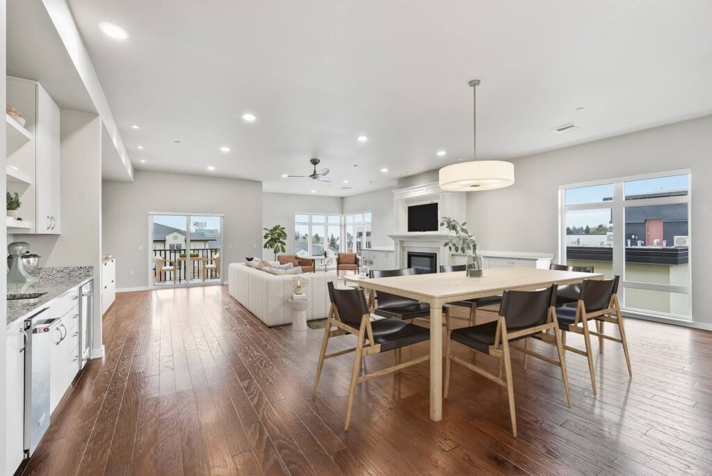 Modern open-concept living and dining area with wood floors, a light wood dining table, black chairs, beige sofa, built-in shelves, large windows, and a fireplace with a wall-mounted TV.