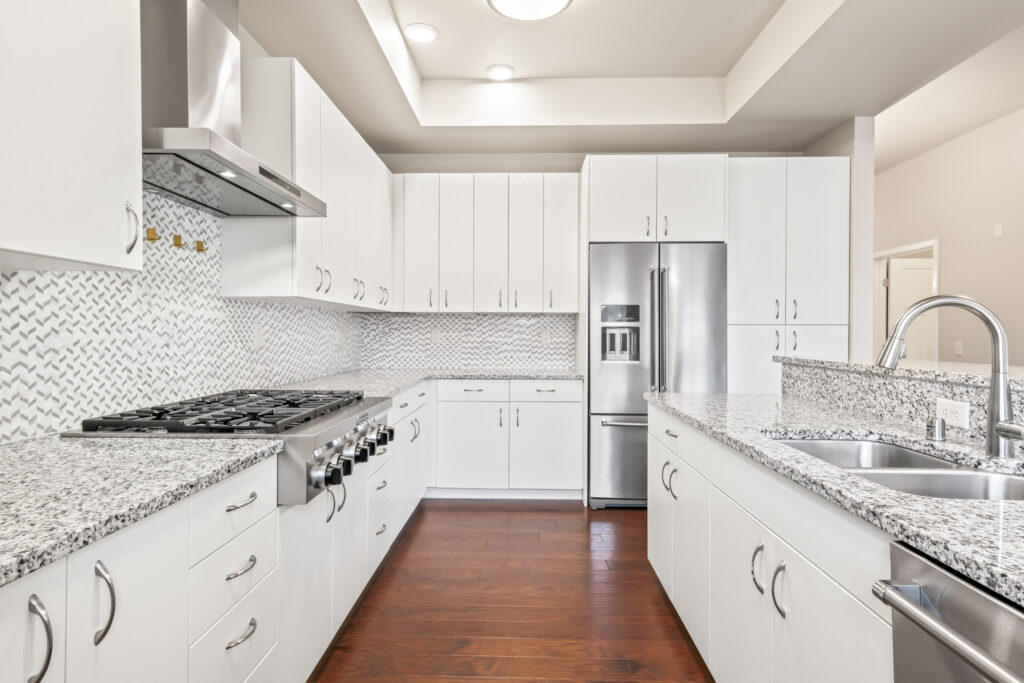 Modern kitchen with white cabinets, stainless steel appliances, granite countertops, gas stove, double sink, and a herringbone tile backsplash, all set on dark wood flooring.
