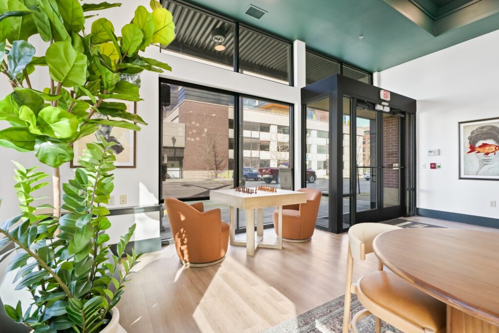 Bright room with large windows, a chessboard on a table between two orange chairs, a large potted plant, and a round wooden table. Art pieces hang on the walls.