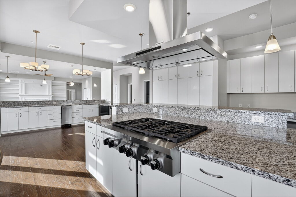 Modern kitchen with white cabinets, granite countertops, a central gas stove with stainless steel range hood, pendant lights, hardwood floors, and an open layout leading to other rooms.