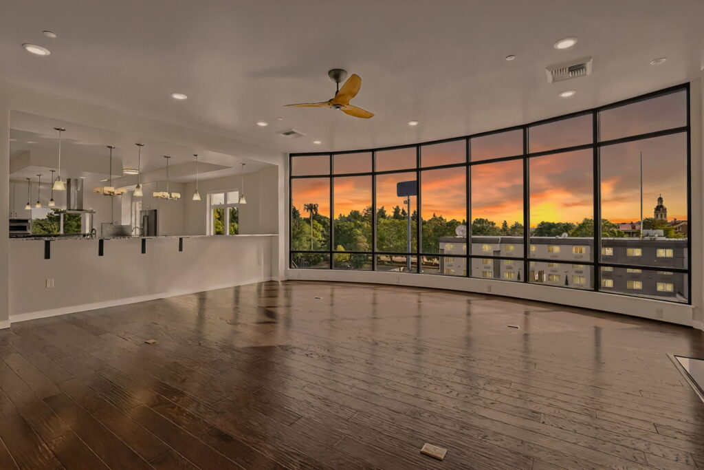 Spacious modern living room with dark wood floors, large curved floor-to-ceiling windows, and a kitchen with hanging lights. The sunset outside shows an orange sky above treetops and distant buildings.