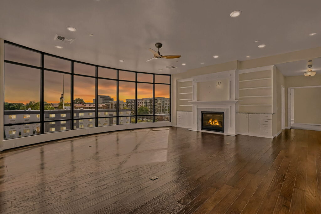 Spacious, empty living room with dark wood floors, large curved floor-to-ceiling windows overlooking a cityscape at sunset, built-in shelves, a modern fireplace, and ceiling fans.