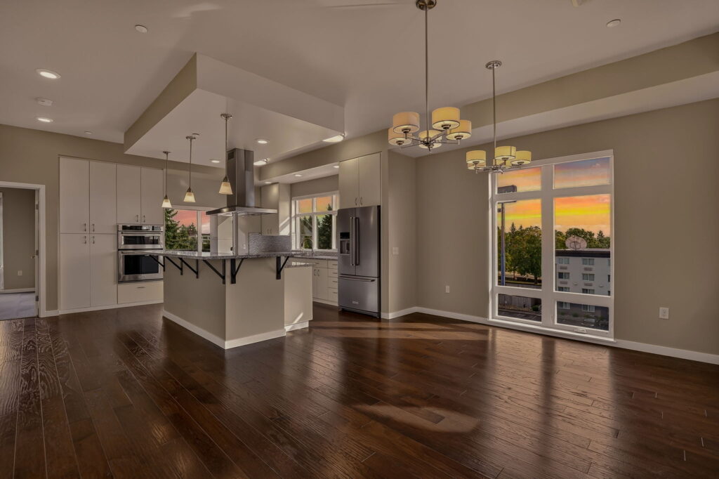 Modern, open-concept kitchen and living area with dark wood floors, white cabinets, stainless steel appliances, pendant lights, and large windows showing a sunset view. Room is empty and well-lit.