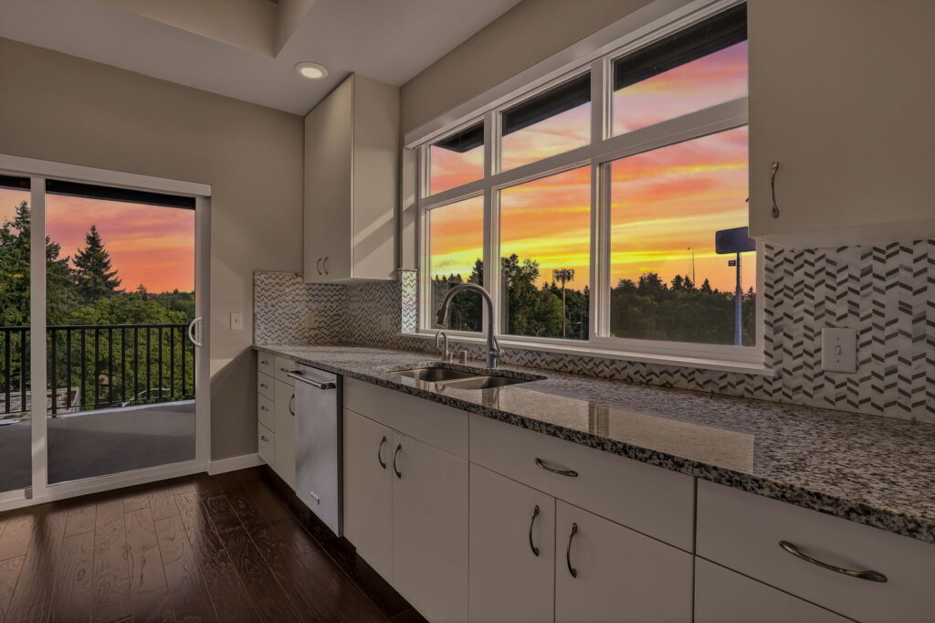 Modern kitchen with white cabinets, granite countertops, and a tile backsplash. Large windows and a sliding glass door reveal a balcony and a vibrant sunset with pink, orange, and yellow hues over a tree-lined view.