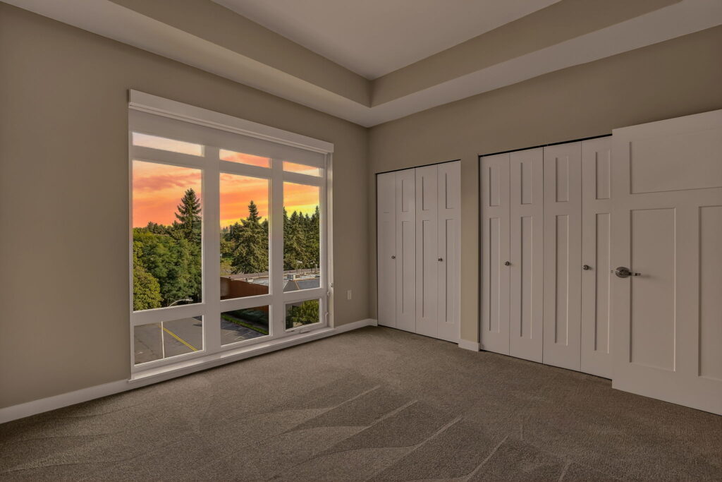 Empty beige-carpeted room with large window overlooking trees and a vibrant sunset, featuring two white double-door closets and neutral-colored walls.