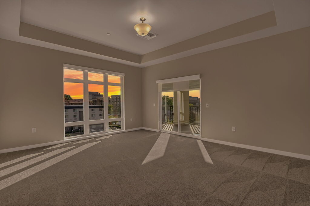 Empty carpeted room with beige walls, a tray ceiling, large windows showing a sunset view, and mirrored closet doors reflecting the outdoor balcony area.