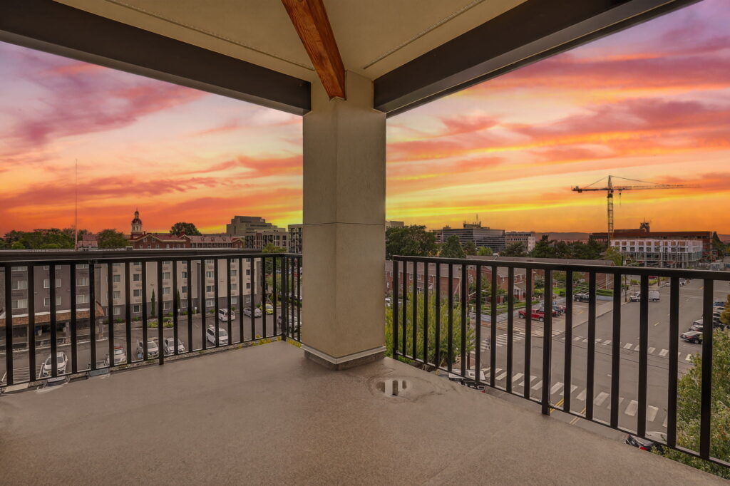 Spacious balcony with black railing overlooking a cityscape at sunset, with vibrant orange and pink clouds, buildings, a crane, trees, and a parking lot in view.