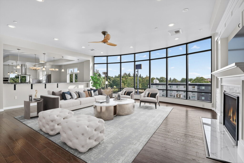 Spacious modern living room with large curved windows, light gray sofas, tufted ottomans, a fireplace, hardwood floors, and views of trees and buildings outside. Contemporary decor and open layout with dining area visible.
