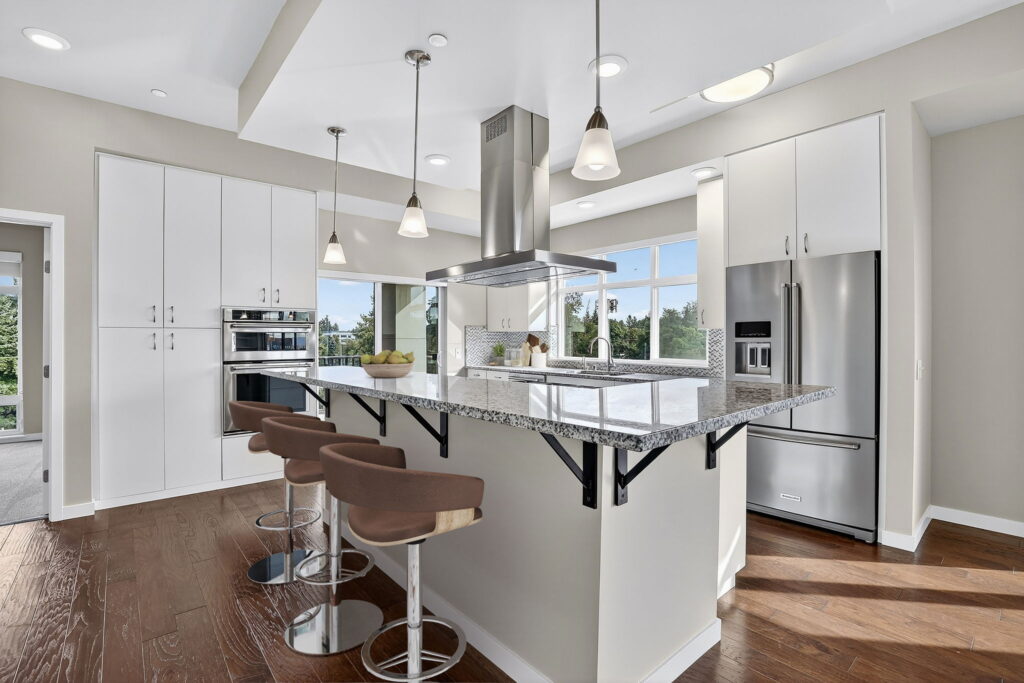 Modern kitchen with white cabinets, stainless steel appliances, granite countertops, three brown bar stools, pendant lighting, hardwood floors, and large windows letting in natural light.