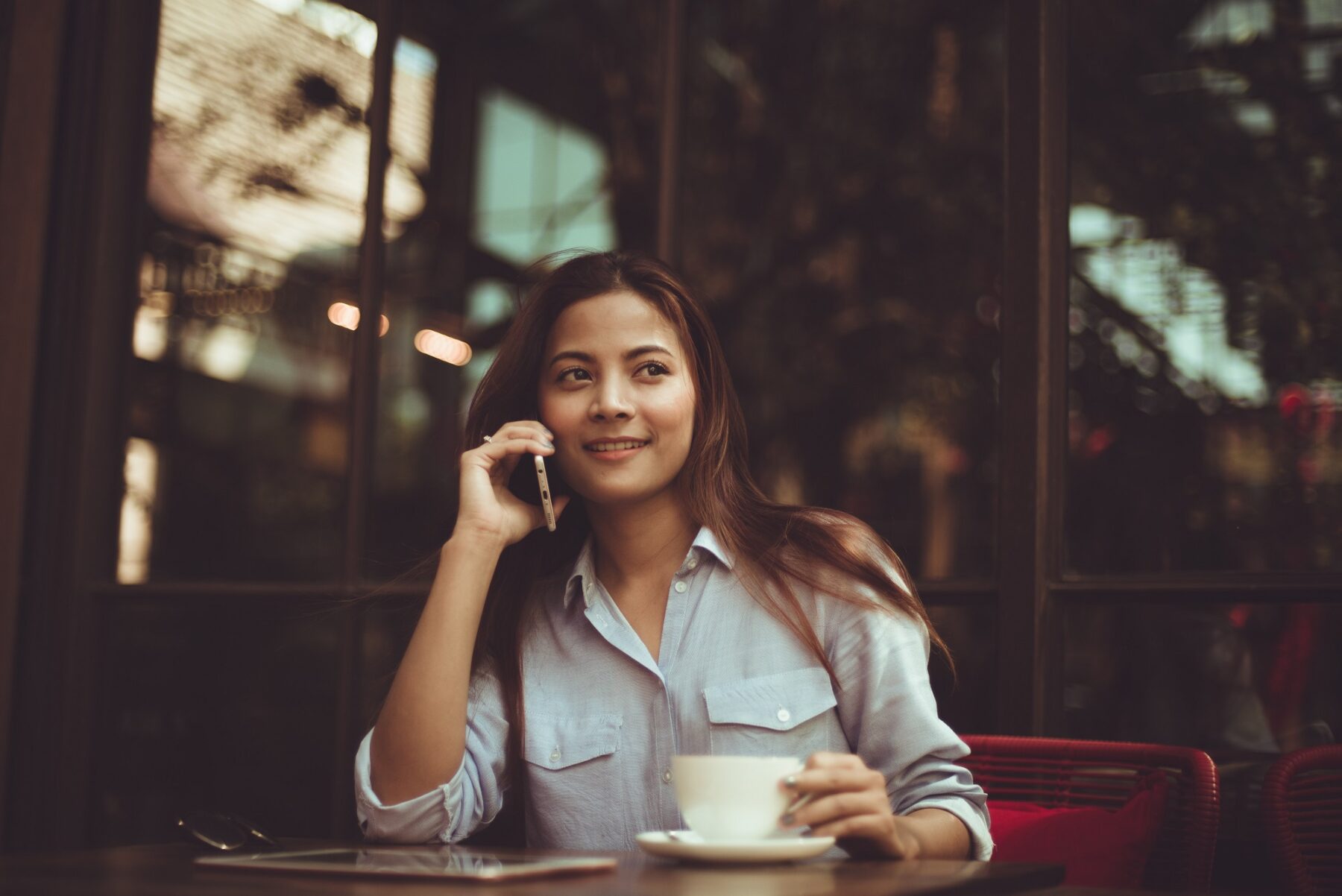 A woman sits at a café table, smiling while talking on her phone and holding a cup of coffee. She wears a light blue shirt, and the background shows large windows with reflections and soft, natural lighting.